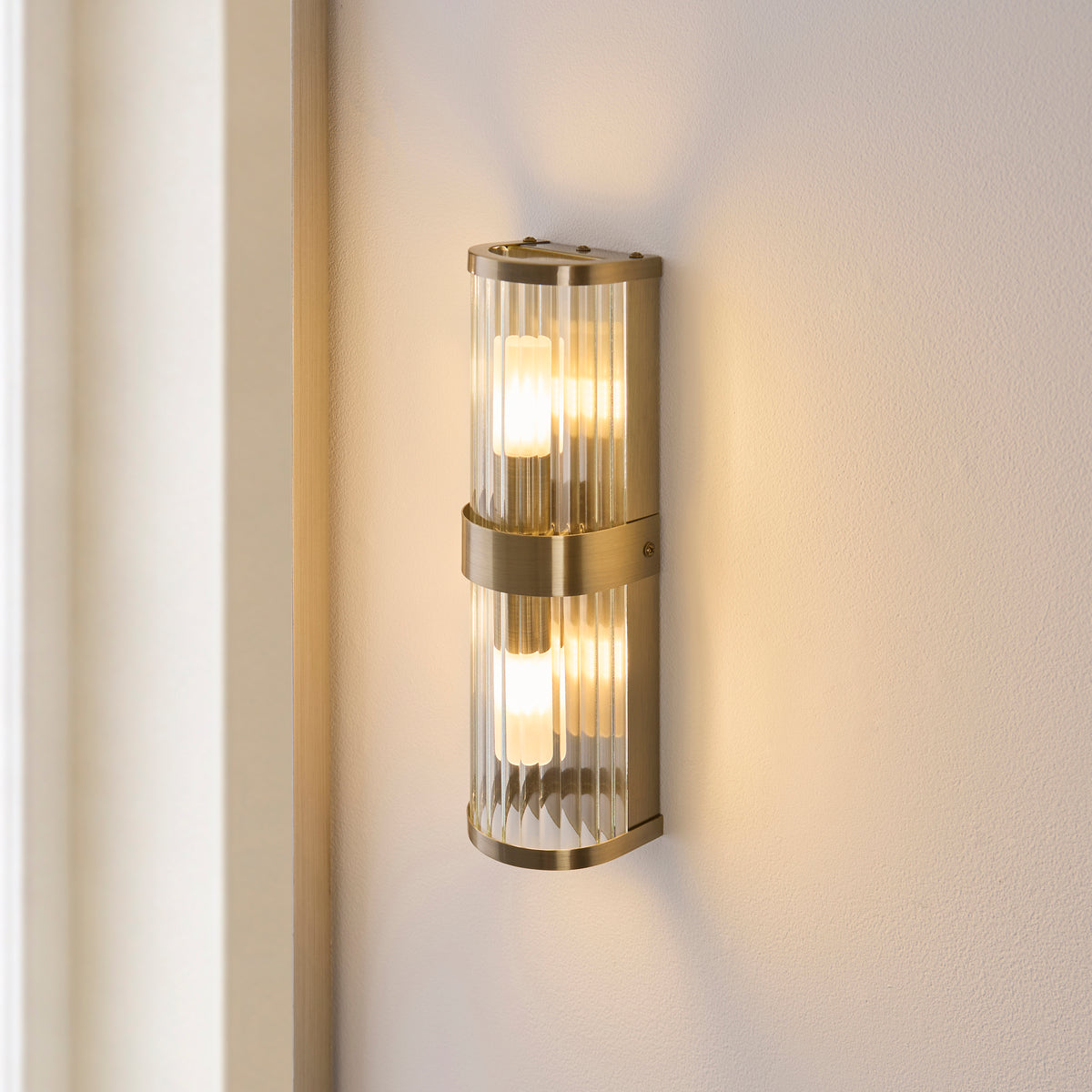 Ridgeton ribbed glass wall light lit on a pale bathroom wall beside a mirror edge