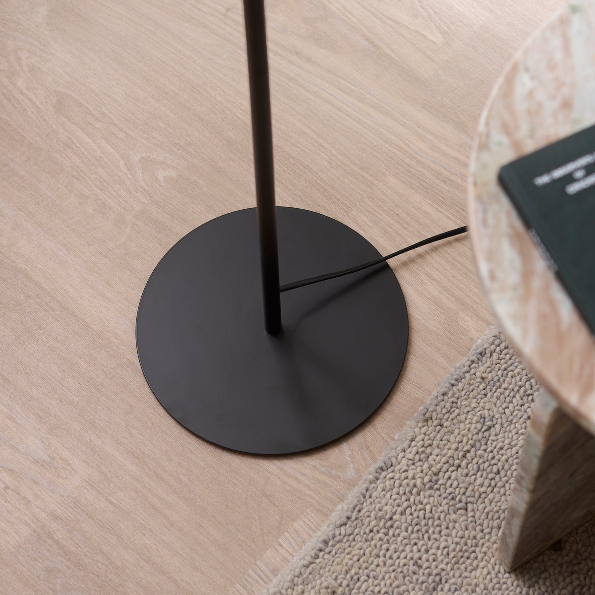 Round black base and stem of the Lyon floor lamp on oak flooring beside a woven rug