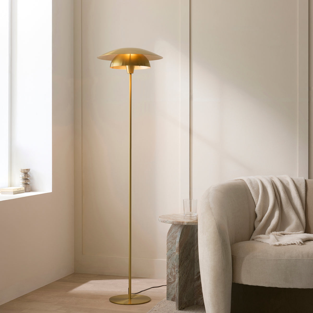Lyon brass floor lamp lit beside a cream sofa and marble table by a sunlit window