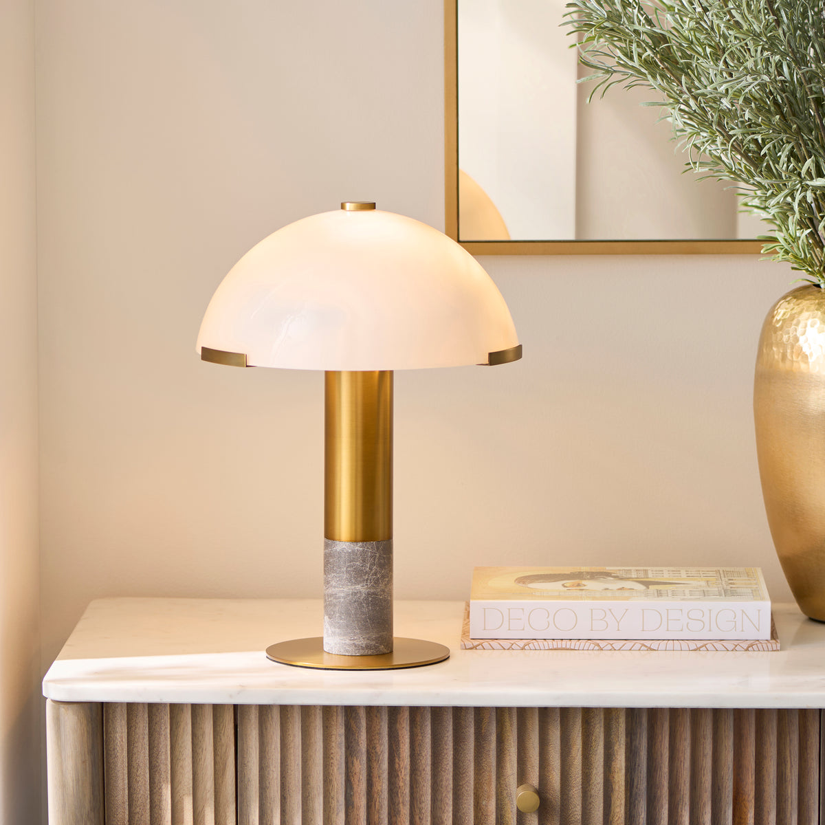 Hansel brass and marble table lamp glowing on a fluted sideboard beside a gold vase and books