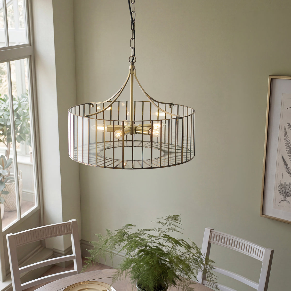 Wide brass Antalya pendant glowing above a dining table beside a bright window