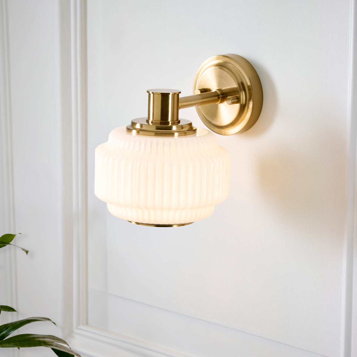 Evora brass wall light beside white panelling, showing the stepped ribbed opal glass shade