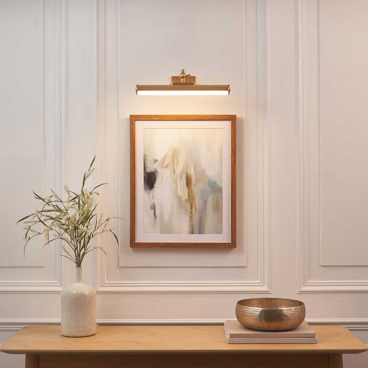 Kindos brass picture light above a framed abstract print on panelled wall, with vase and bowl on console