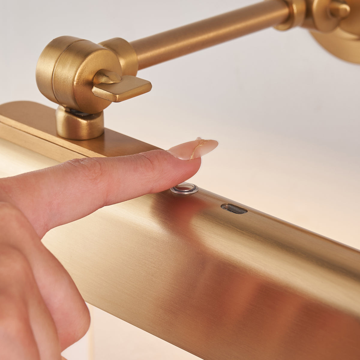 Fingertip pressing the touch dimmer switch on the brass shade of the Dalyan picture light