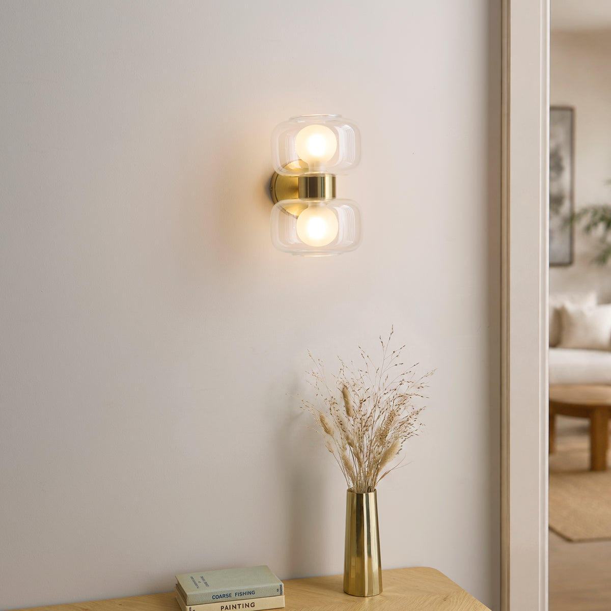 Rossano brass wall light lit beside a console table with dried grasses in a brass vase