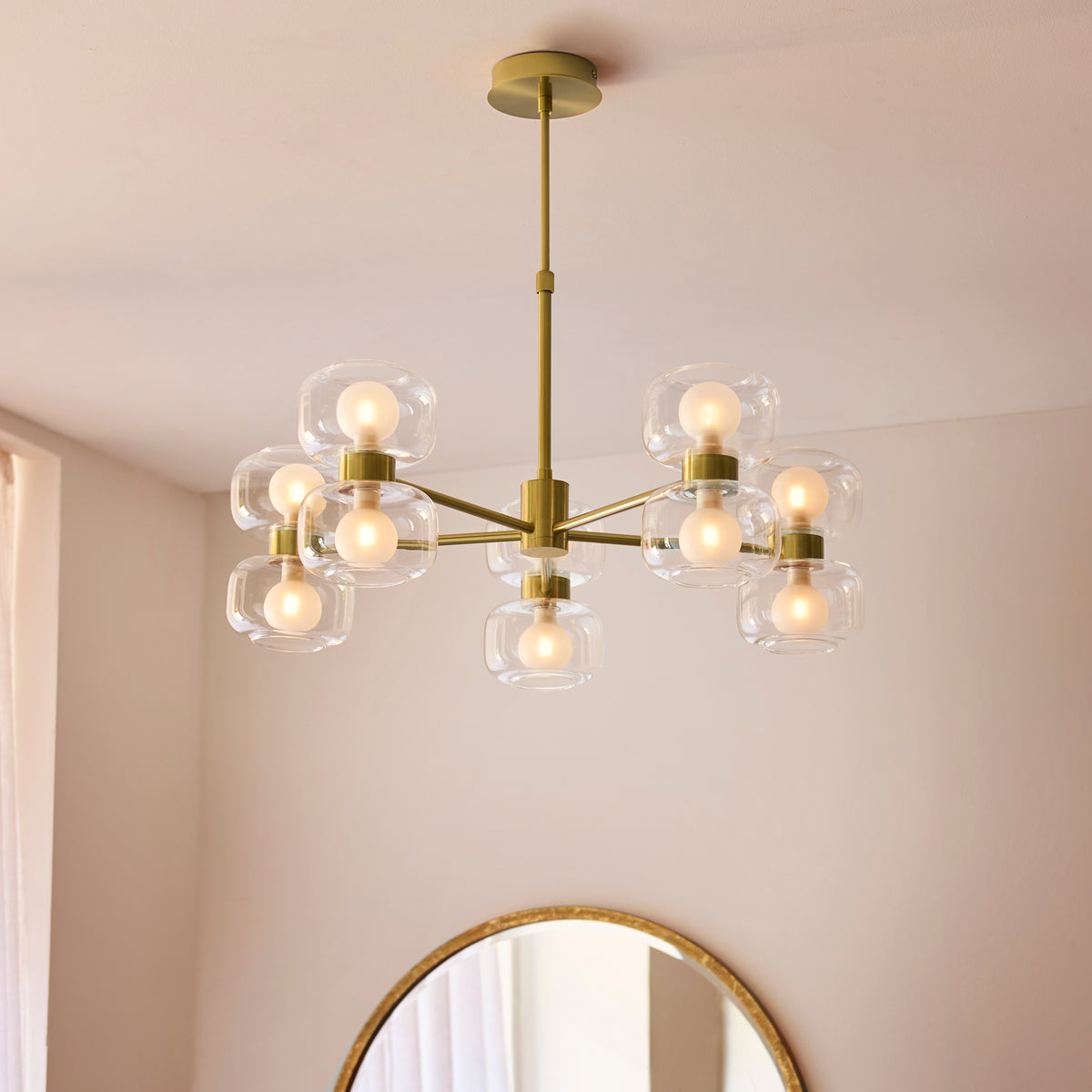 Rossano brass chandelier lit in a bedroom above a round mirror and pale curtains