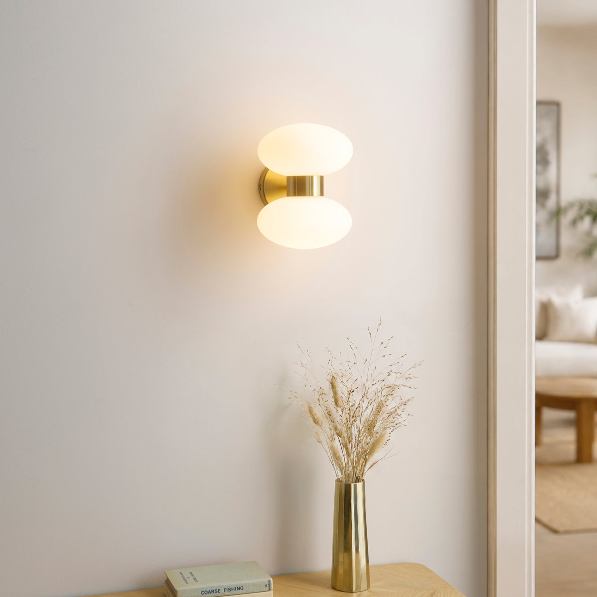 Rossano brass wall light lit above a hallway console with brass vase of dried grasses and books
