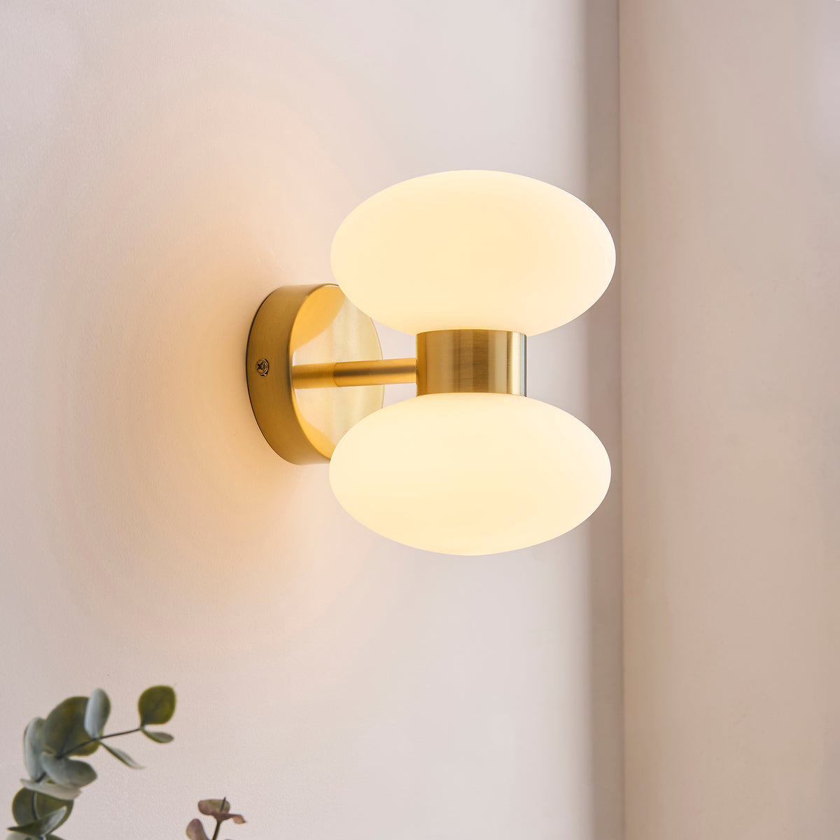 Rossano brass wall light glowing on a pale corner wall beside eucalyptus foliage