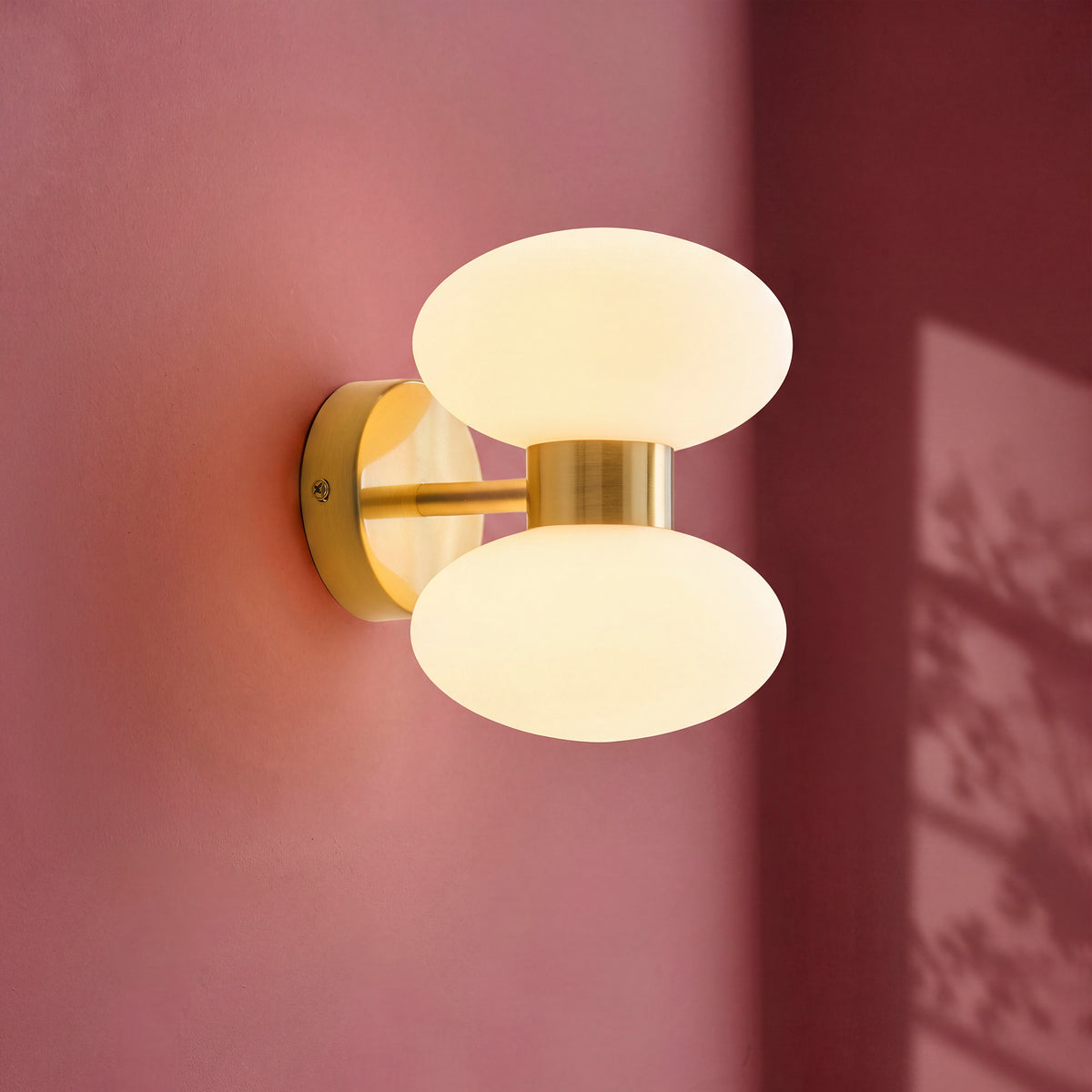 Rossano brass twin opal glass wall light lit against a dusky pink wall with leaf shadows