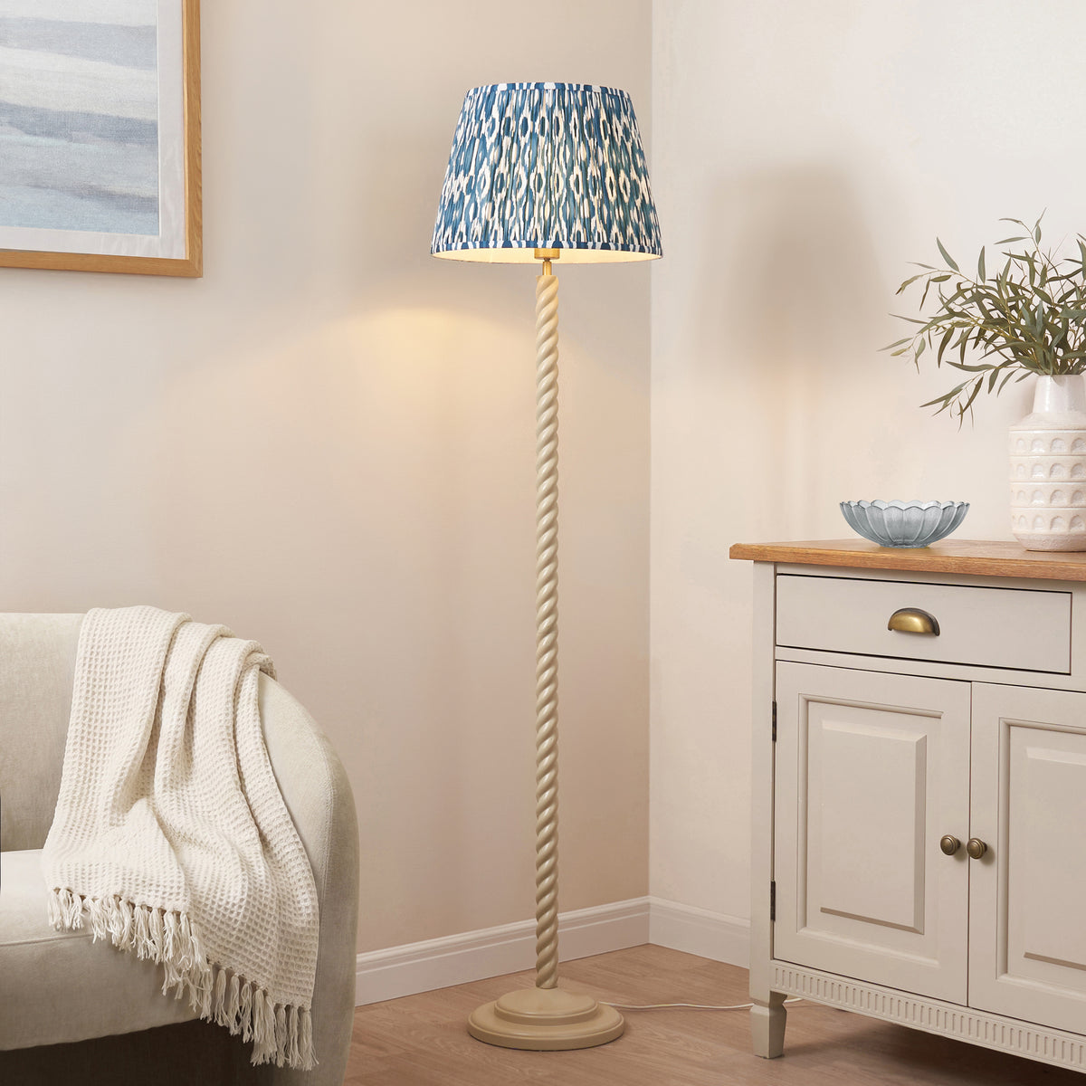 Blue ikat shade floor lamp lit in a living room corner beside a cream sideboard and armchair with throw