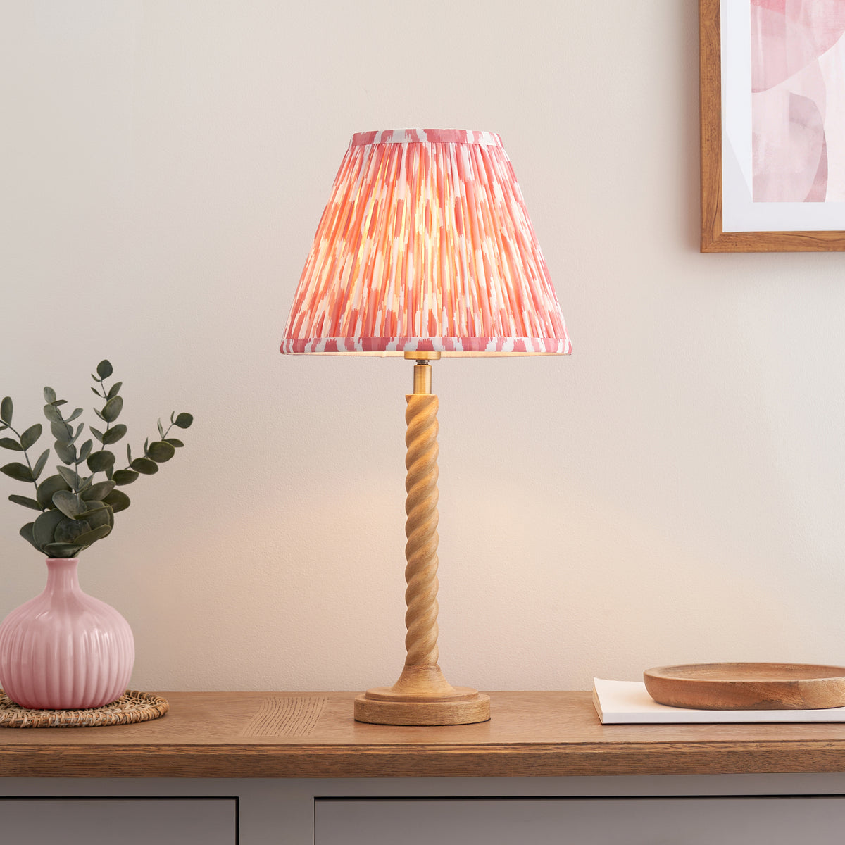 Coral pink ikat table lamp lit on an oak sideboard beside a pink vase with eucalyptus and a wooden dish