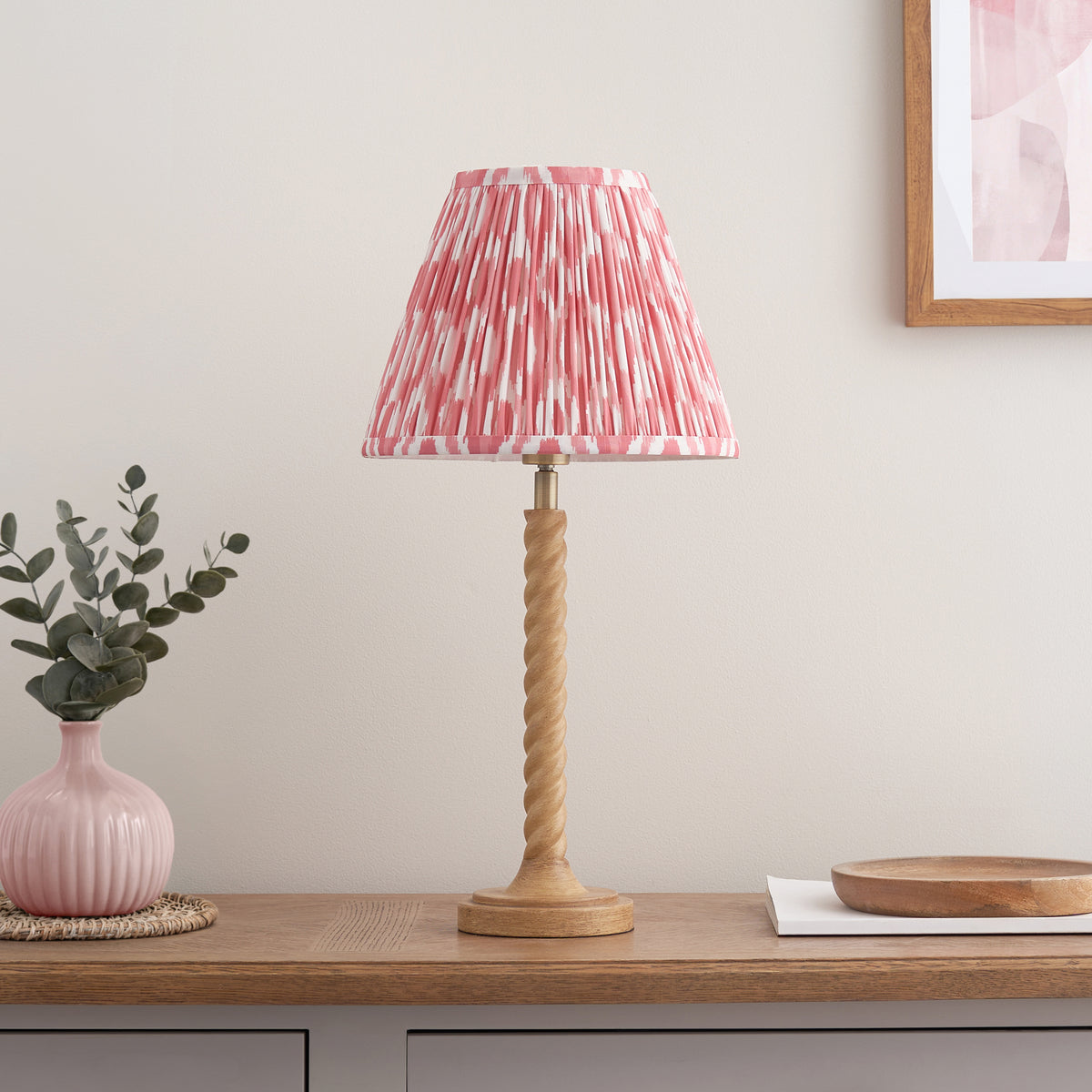 Oak effect twisted table lamp with coral pink ikat shade styled on a wooden console below abstract art