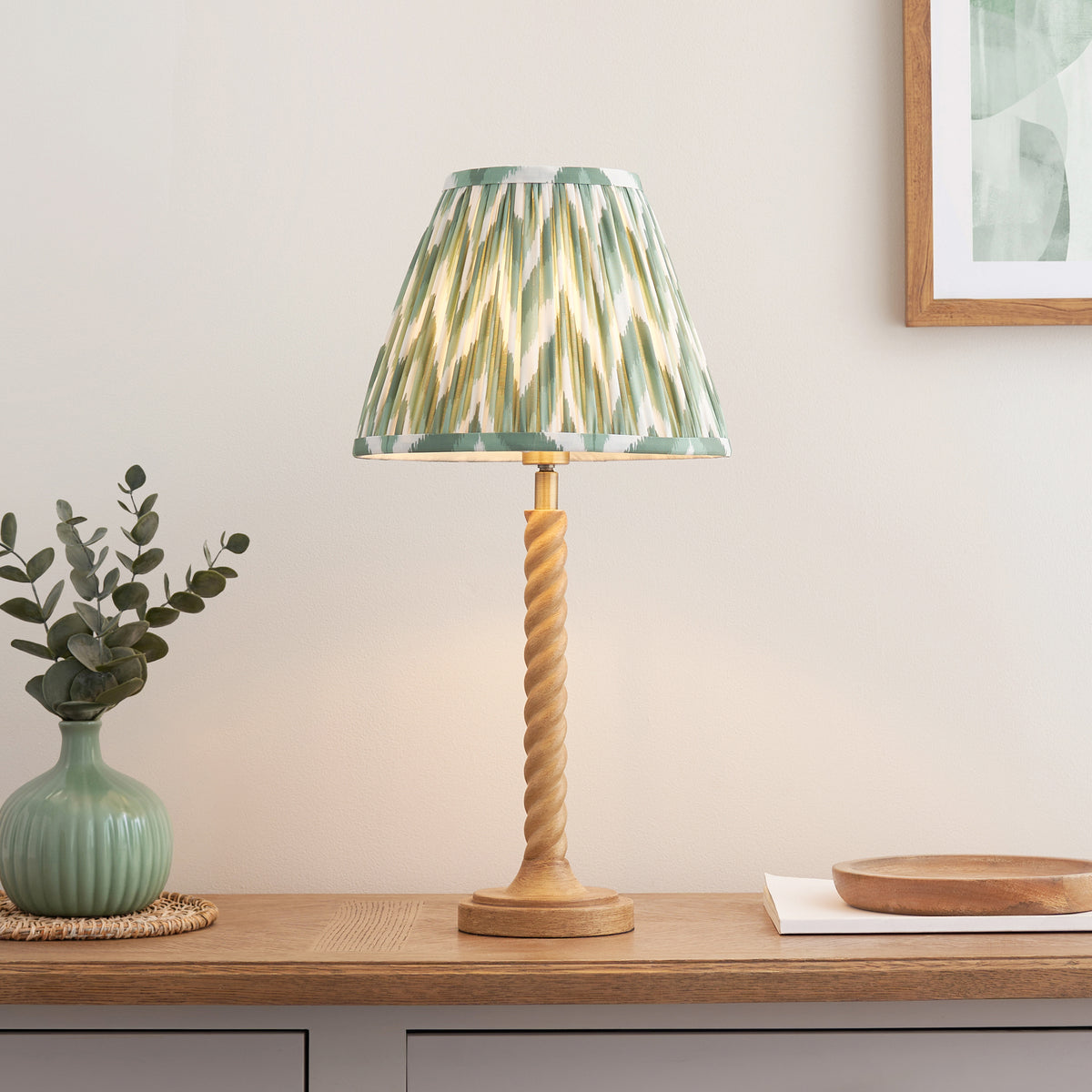 Green zigzag shade table lamp lit on an oak sideboard beside a green vase of eucalyptus