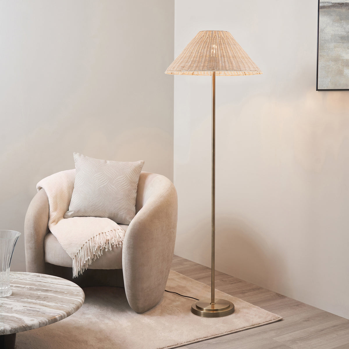 Arlo antique brass floor lamp with rattan shade lit beside a cream boucle armchair in a neutral living room