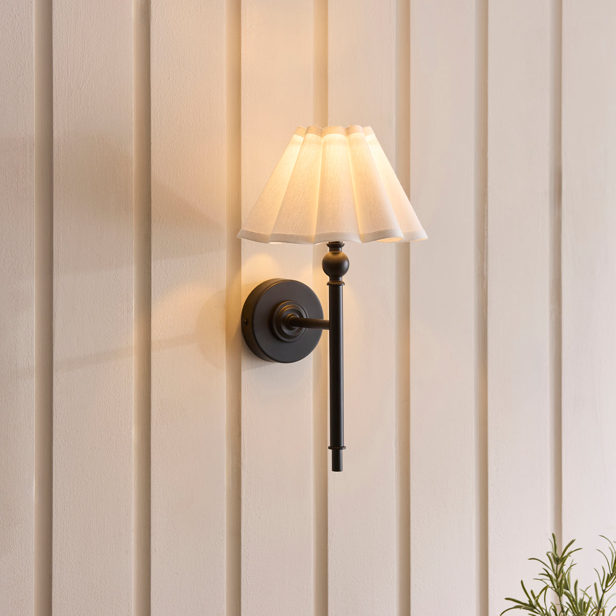 Sylvie matt black wall light glowing against cream panelled walls beside a potted rosemary plant
