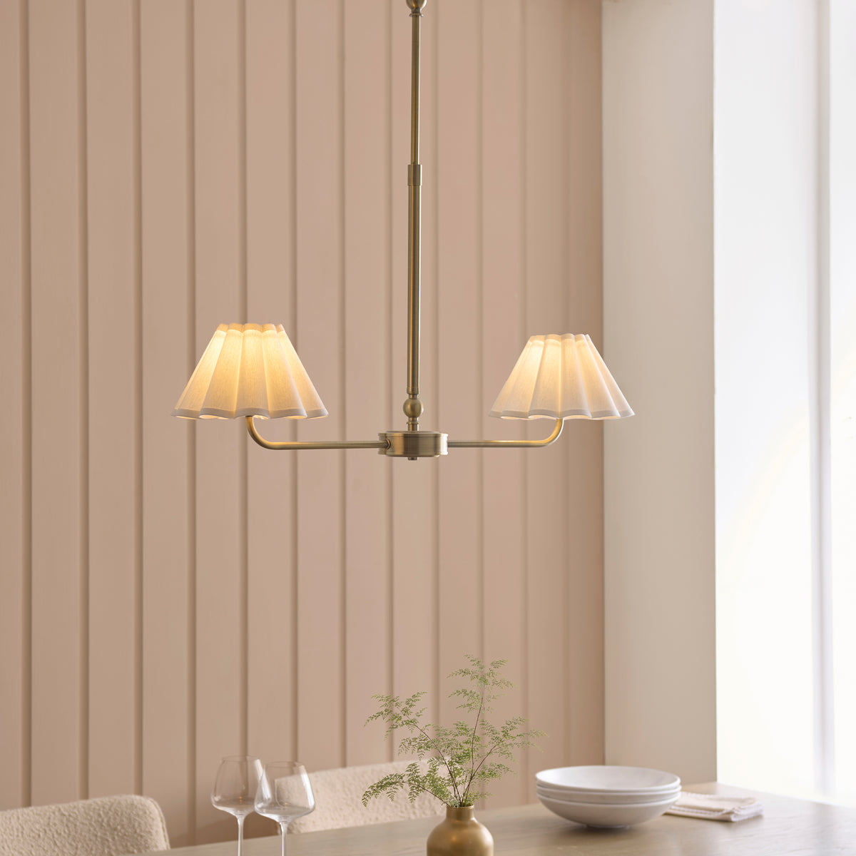 Sylvie brass linear pendant lit above a dining table with bowls, glasses and a vase of foliage