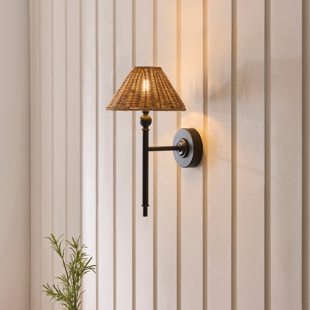 Jericho black wall light lit on a cream panelled wall above a small potted rosemary plant