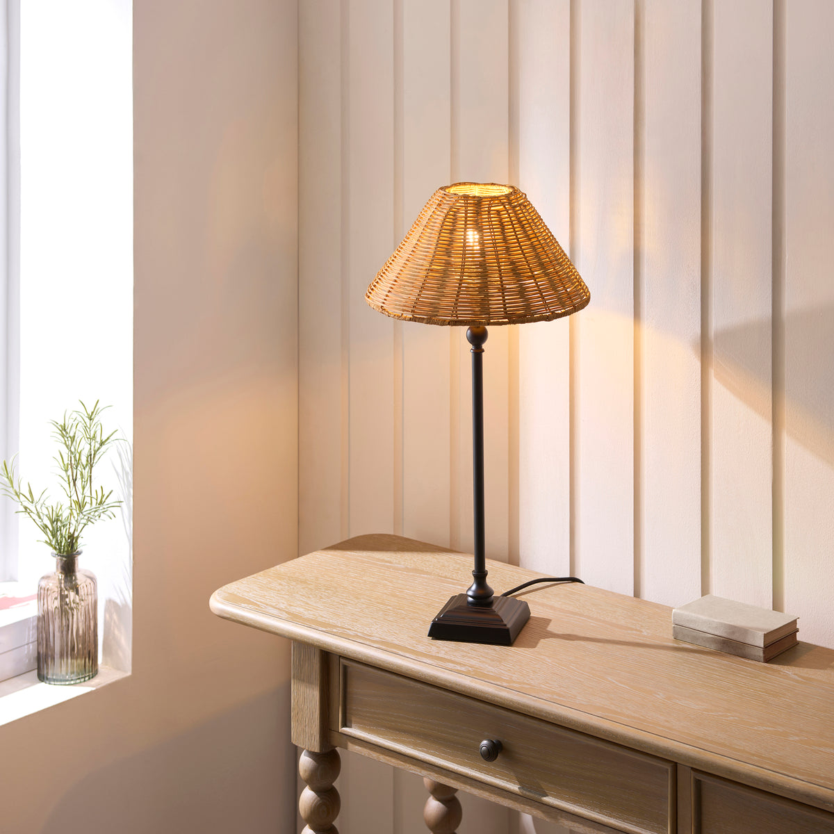 Jericho black table lamp lit on a wooden console table beside a window and glass vase