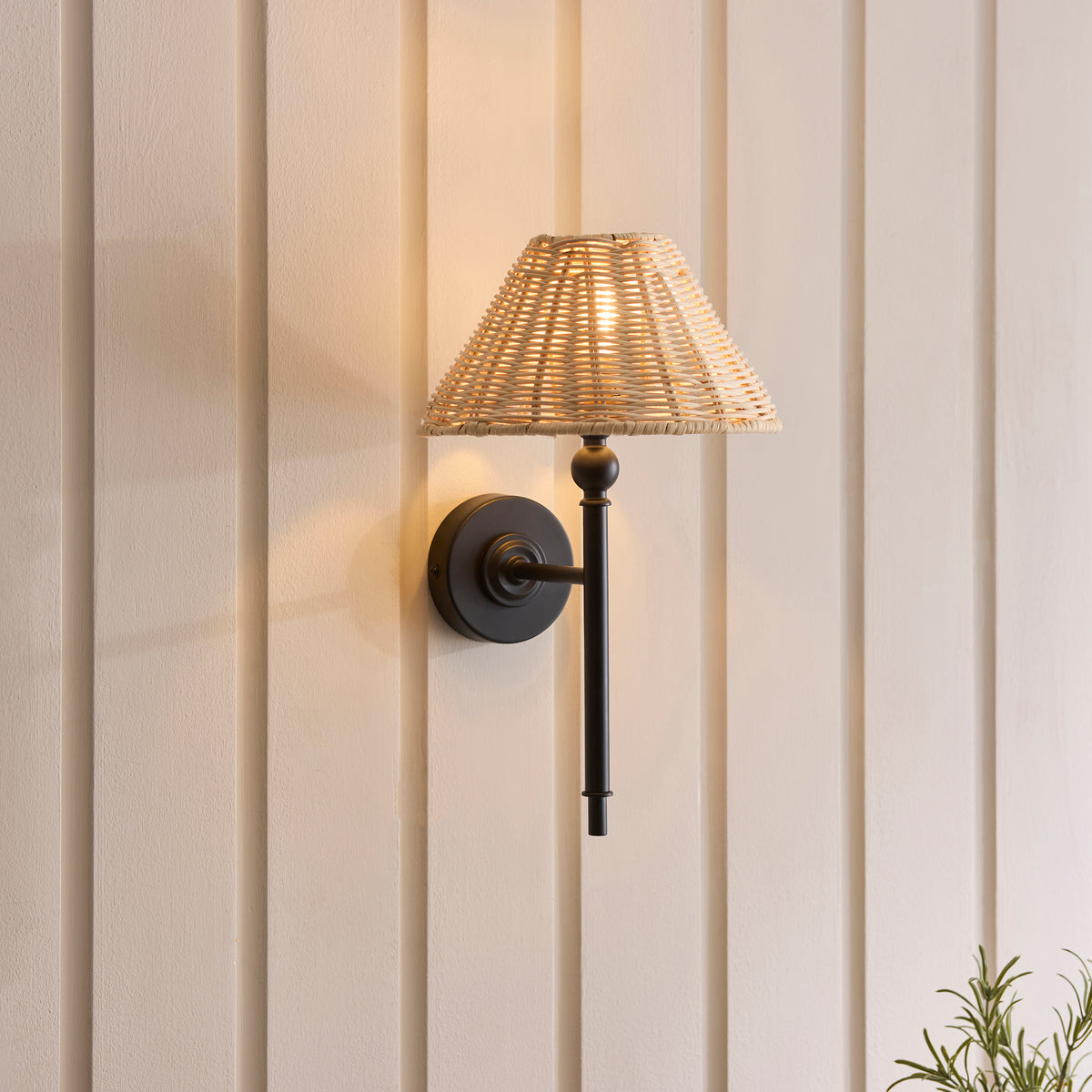 Jericho black wall light lit on cream panelling beside a small potted rosemary plant