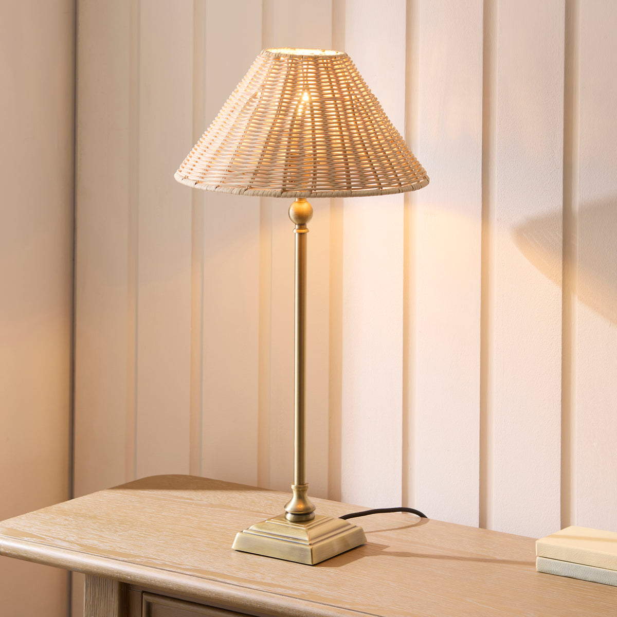 Lit Jericho brass table lamp with rattan coolie shade on an oak sideboard against cream panelling