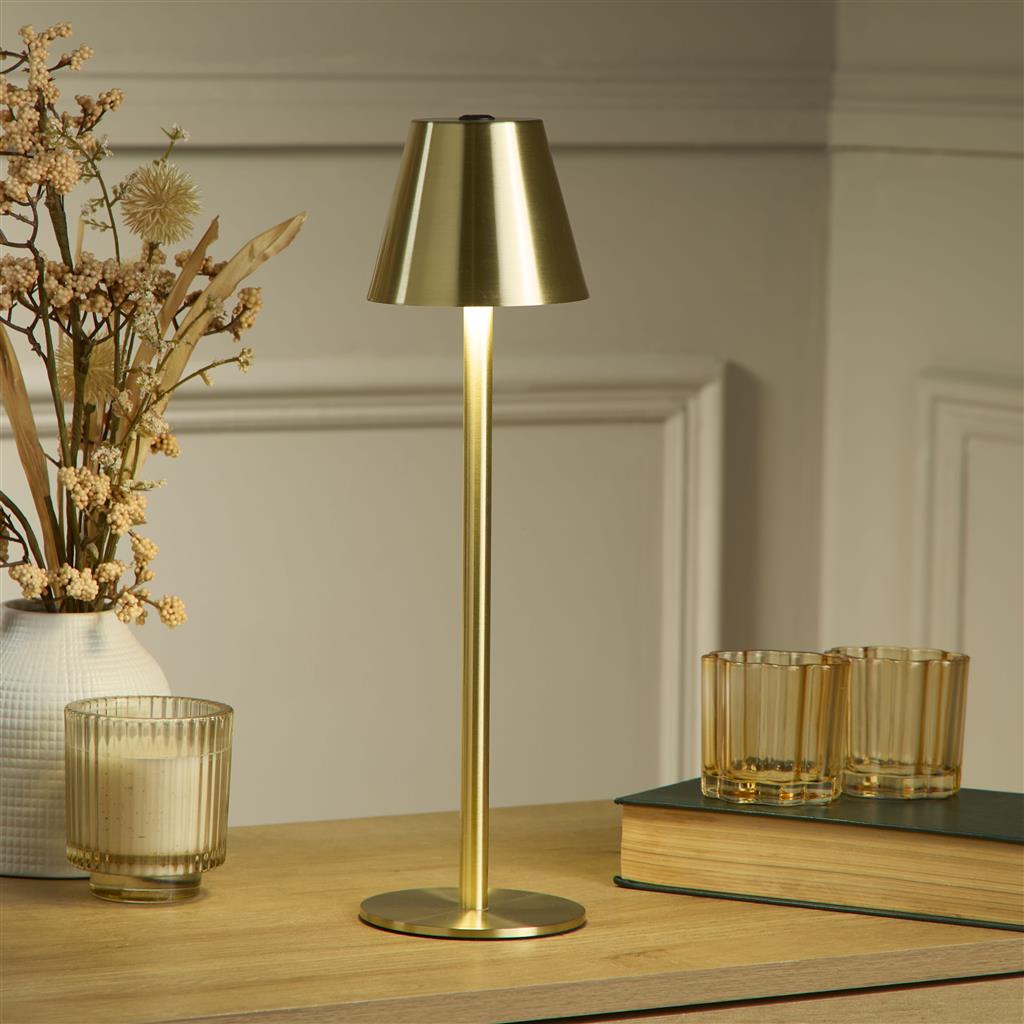 Gold rechargeable table lamp on a wooden surface with decorative items, including books and a vase.