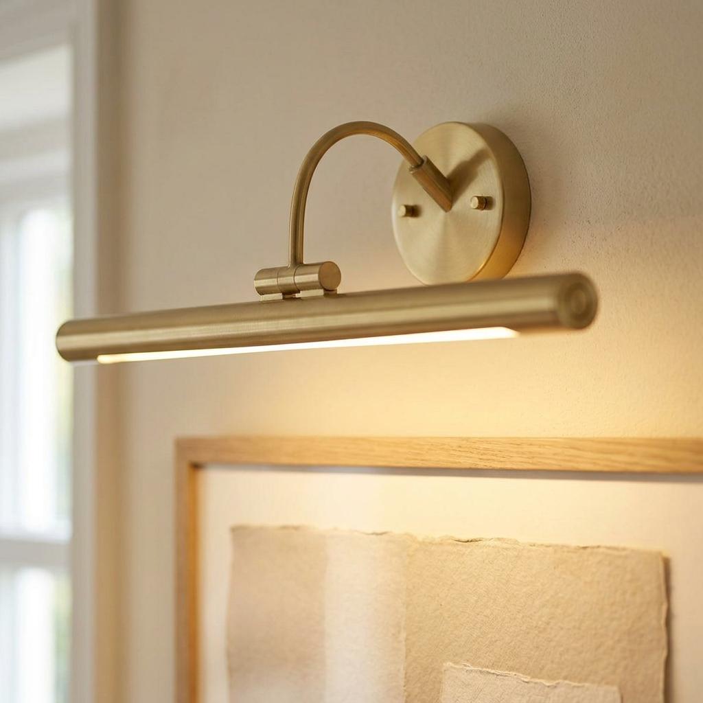 A sleek brushed brass LED picture light mounted on a wall, illuminating a framed piece of artwork below.