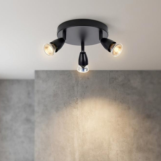 Black three-light round ceiling spotlight illuminating a minimalist room with a concrete wall and modern chair.