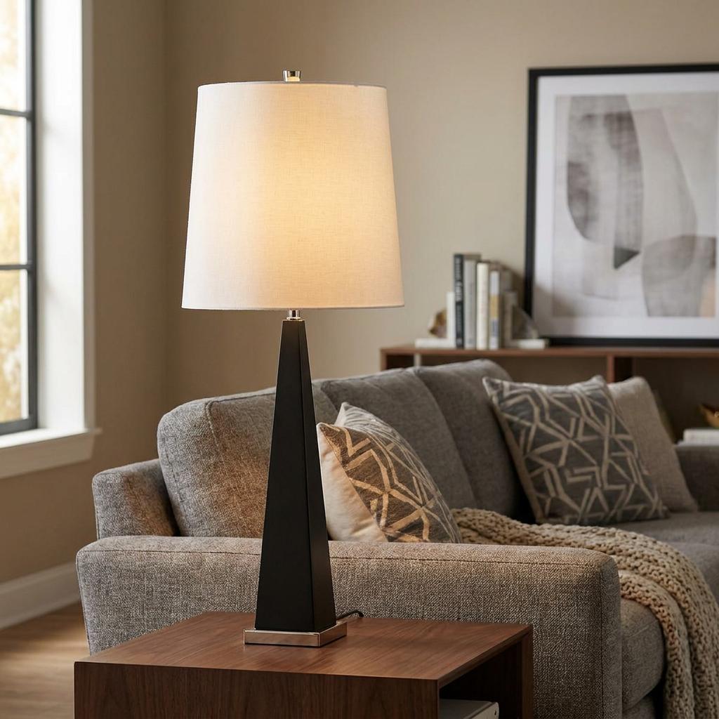 Modern black table lamp with a tapered base and white shade lit on a wooden table beside a grey sofa.