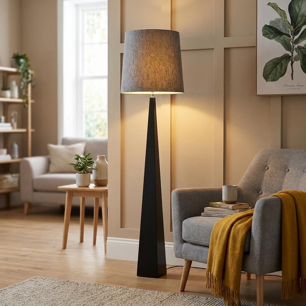 Modern black tapered floor lamp with a grey fabric shade standing next to a grey armchair in a cozy living room.