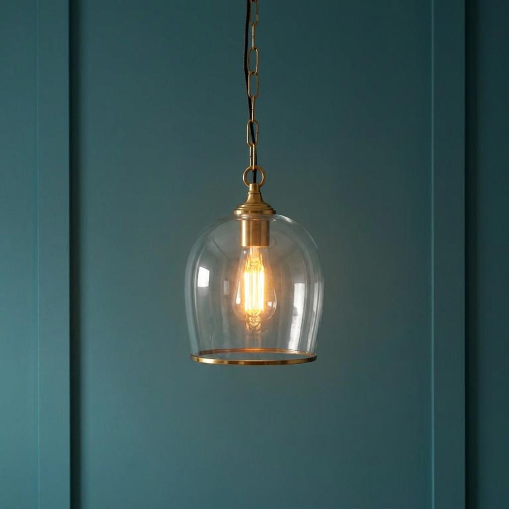 Ashcroft brass ceiling pendant with a clear glass dome shade and lit bulb against a teal paneled wall.