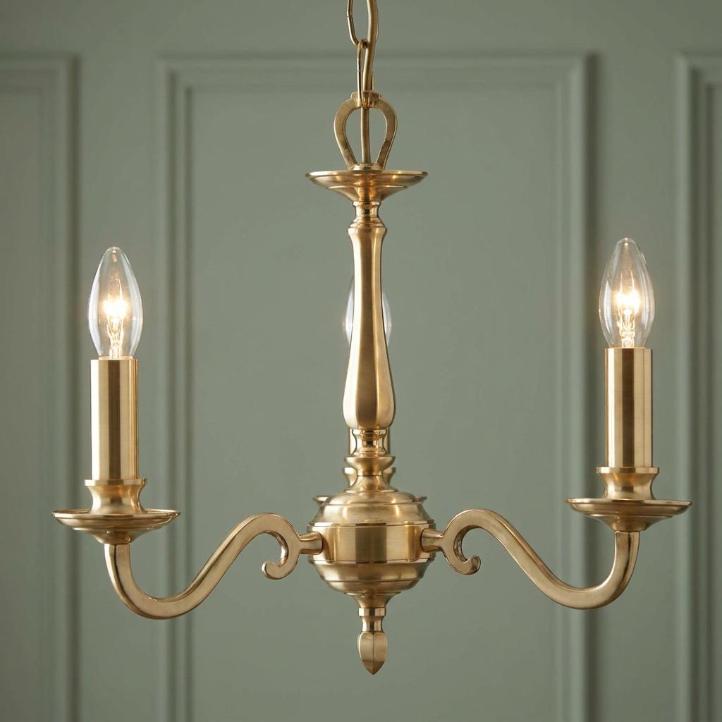 Gold three-light candle-style chandelier with illuminated bulbs against a green paneled wall.