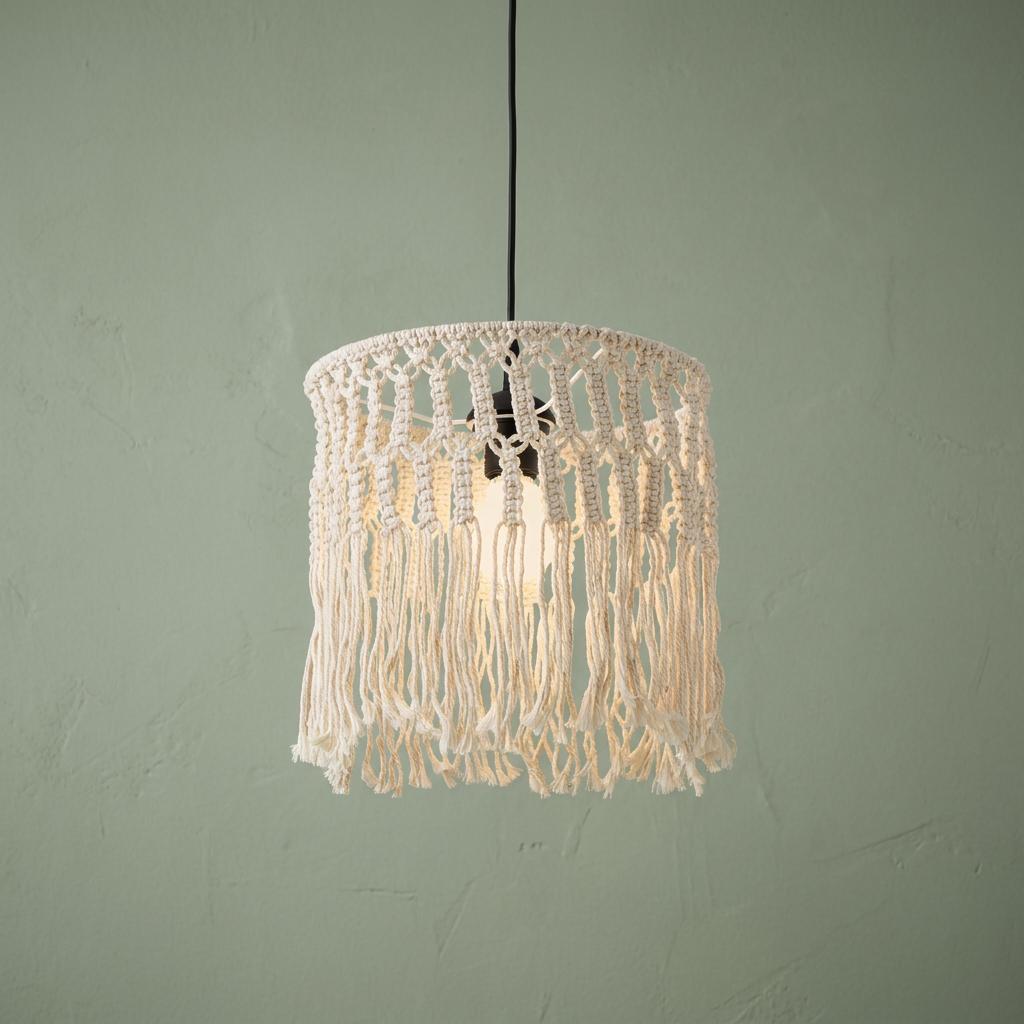 Macrame pendant light with cream fringe and a lit bulb hangs against a textured sage green wall.
