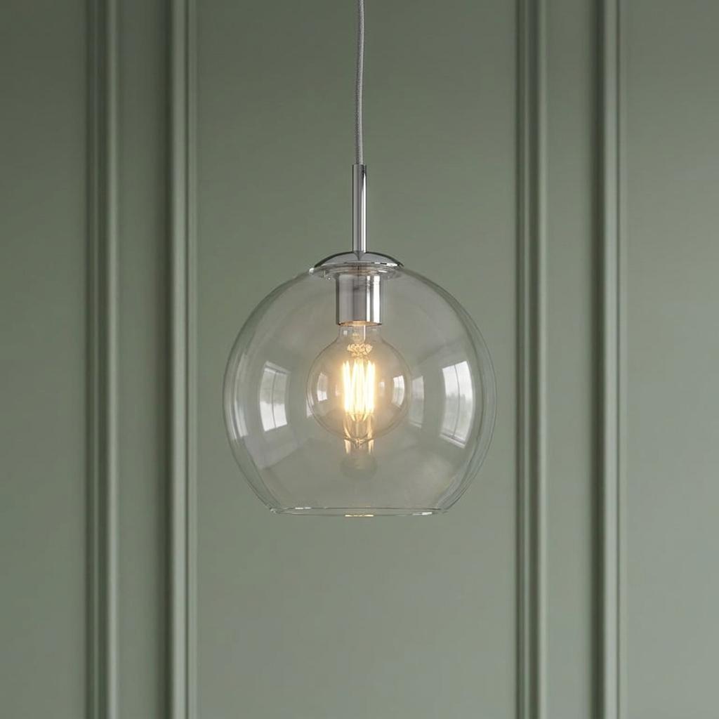 Modern clear glass globe pendant light with a warm glowing filament bulb against a green paneled wall.