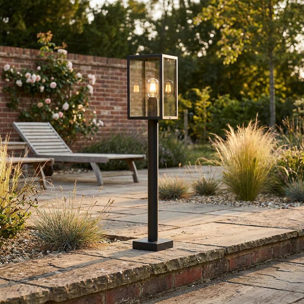 Modern black rectangular pedestal lantern with a glowing bulb on a stone garden patio.