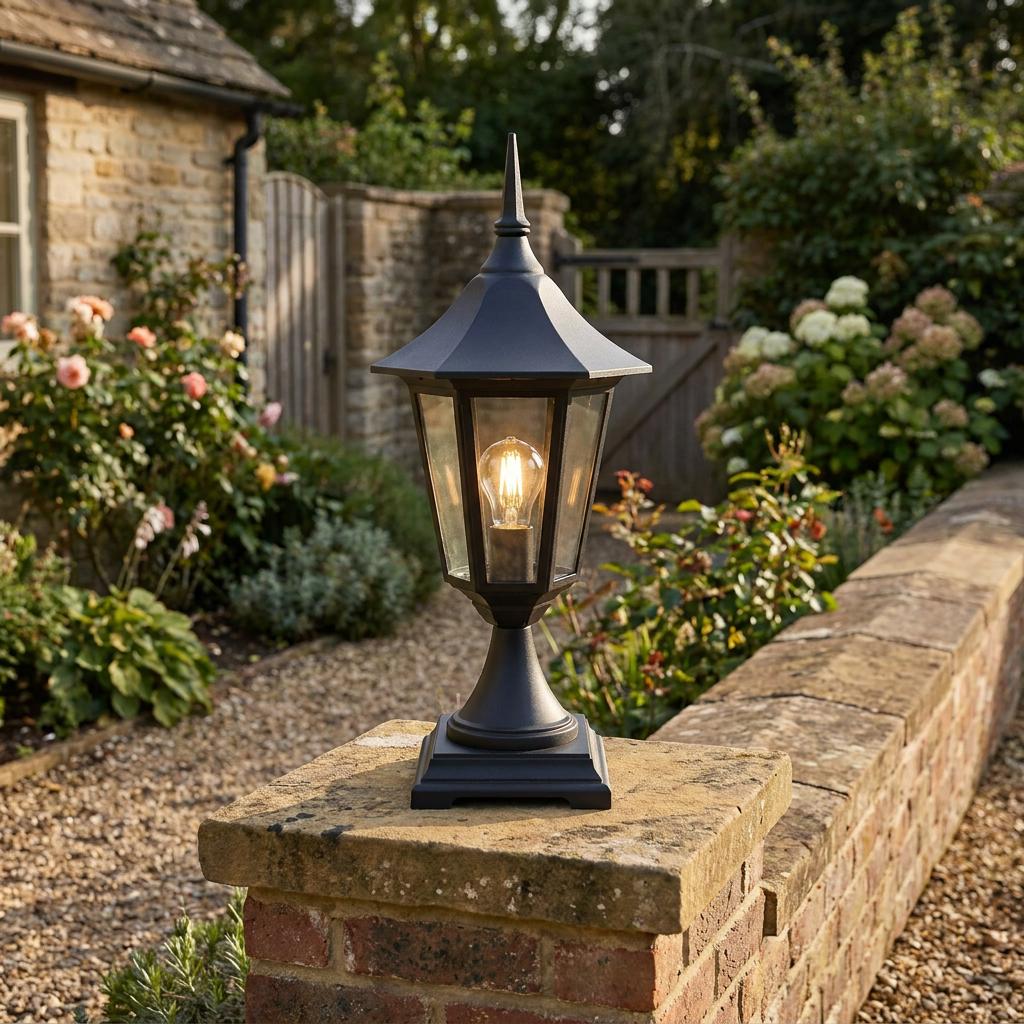Black lantern-style pedestal light with a warm glow, mounted on a stone garden wall pillar in a cottage garden.