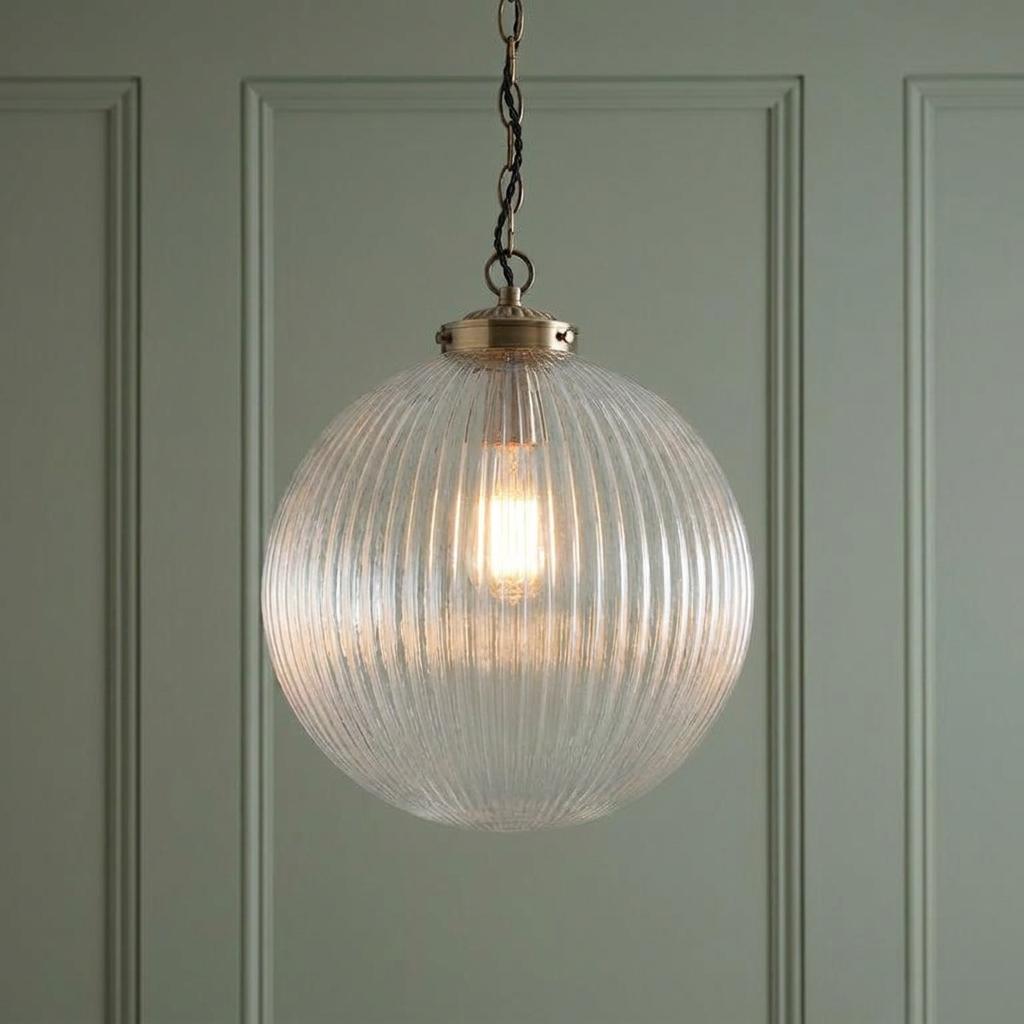 A spherical pendant light with clear ribbed glass and brass fixtures against a pale green paneled wall.