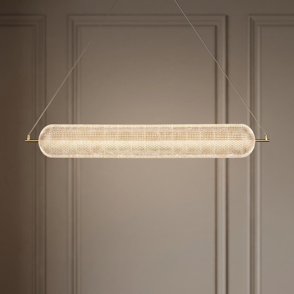 Long capsule-shaped LED pendant with textured acrylic shade and satin brass accents against a neutral paneled wall.