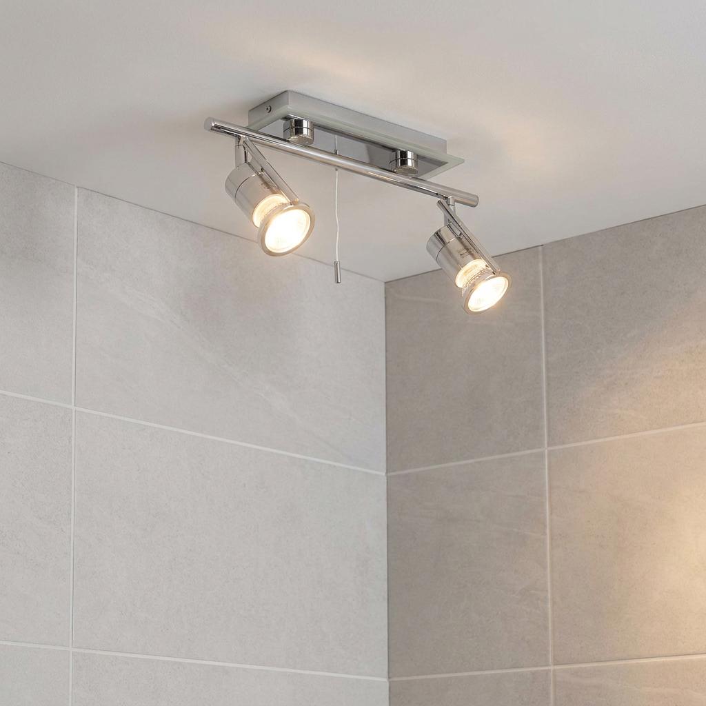 Modern chrome two-light ceiling spotlight with a pull cord mounted in a bathroom with grey wall tiles.