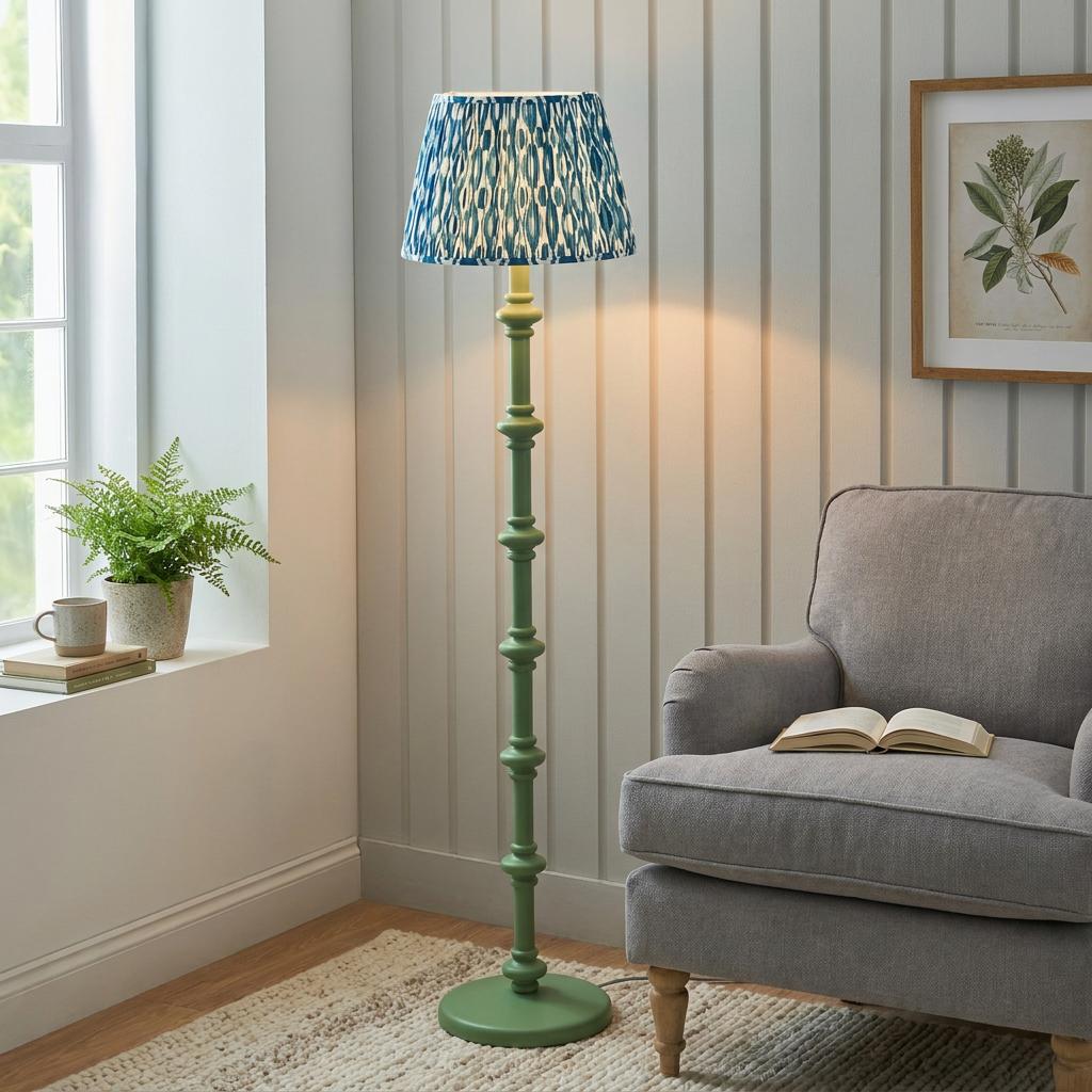 Green wooden floor lamp with a blue ikat pattern shade next to a grey armchair in a wood-paneled room.