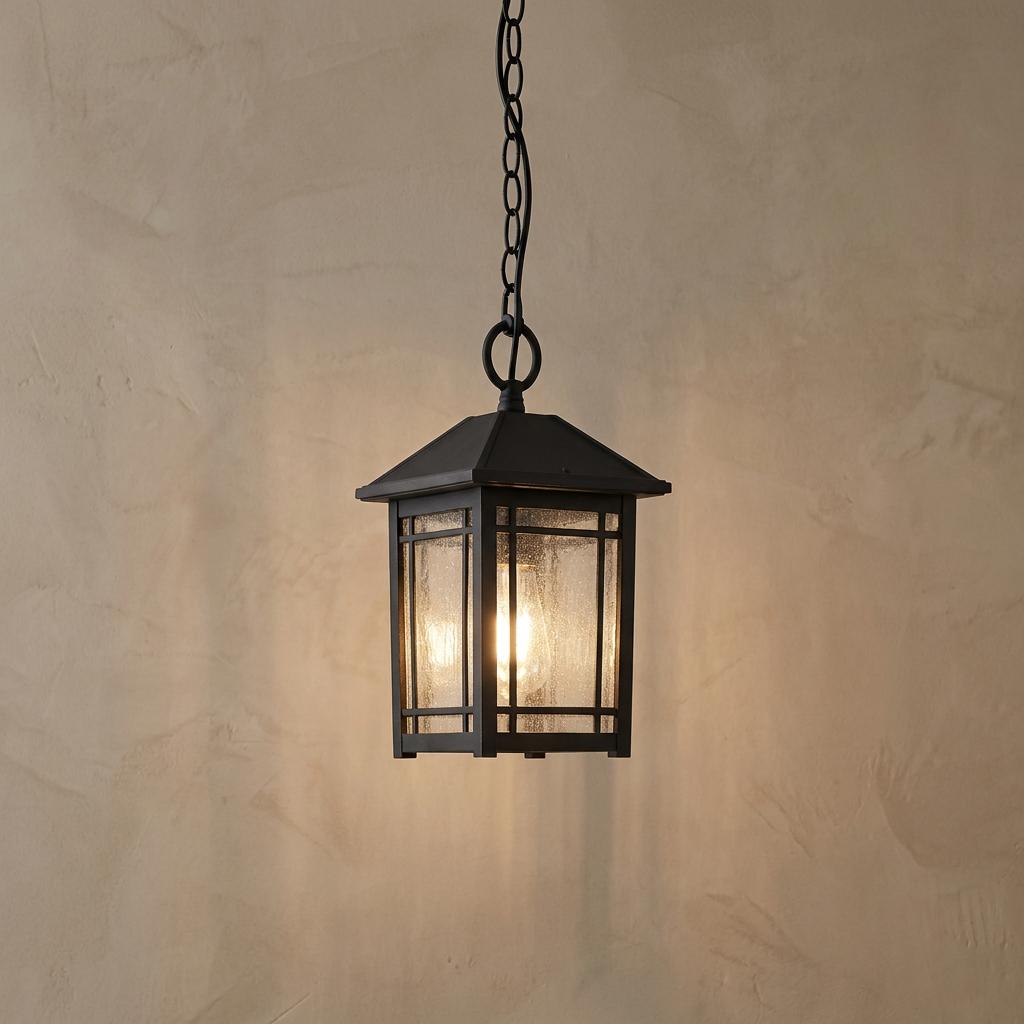 Black outdoor chain lantern with seeded glass panels glowing against a neutral textured wall.
