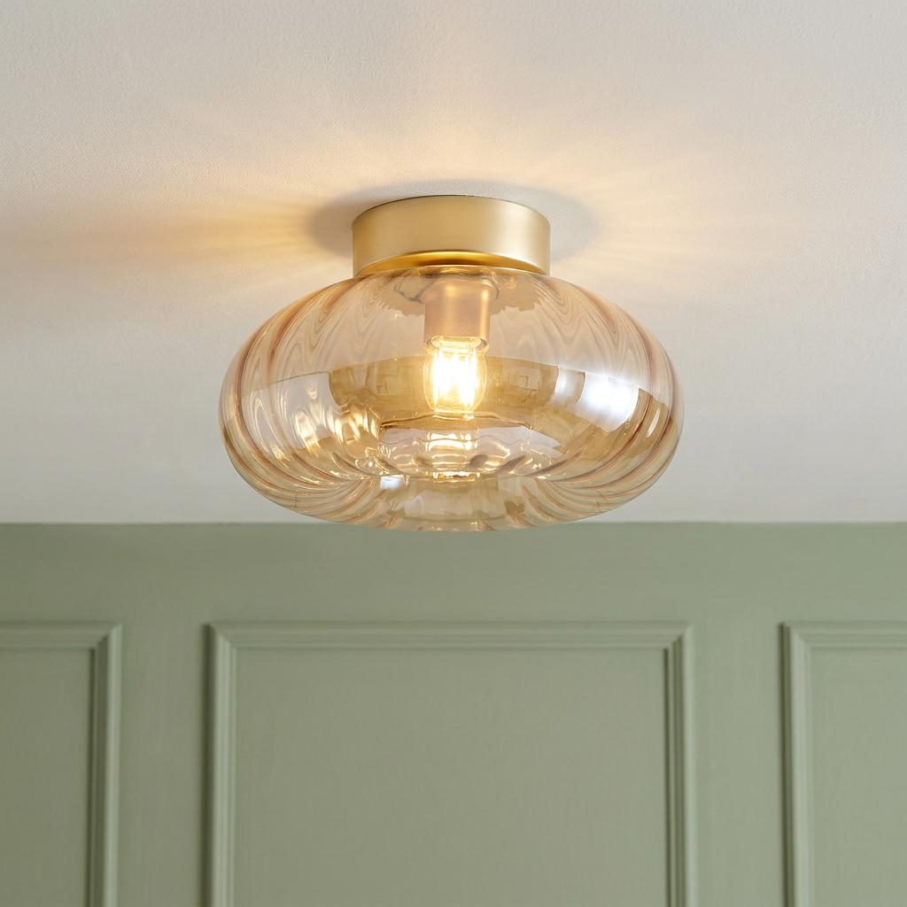 Gold flush ceiling light with a ribbed amber glass shade, illuminated on a white ceiling above a green paneled wall.