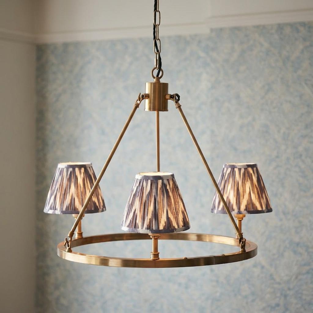 Brass chandelier with three grey zigzag pleated shades set against blue patterned wallpaper.
