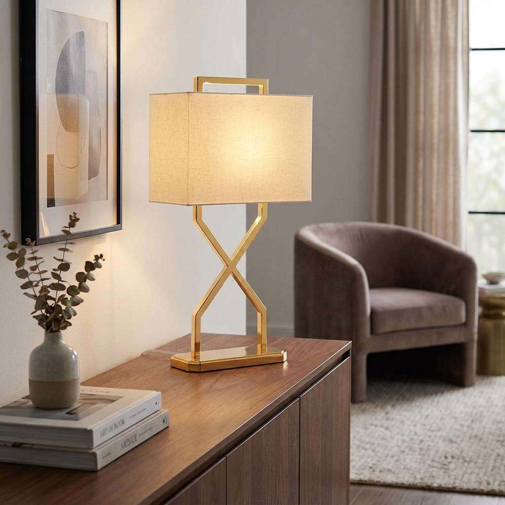 Modern gold table lamp with a cross-base and ivory rectangular shade on a wooden sideboard in a living room.