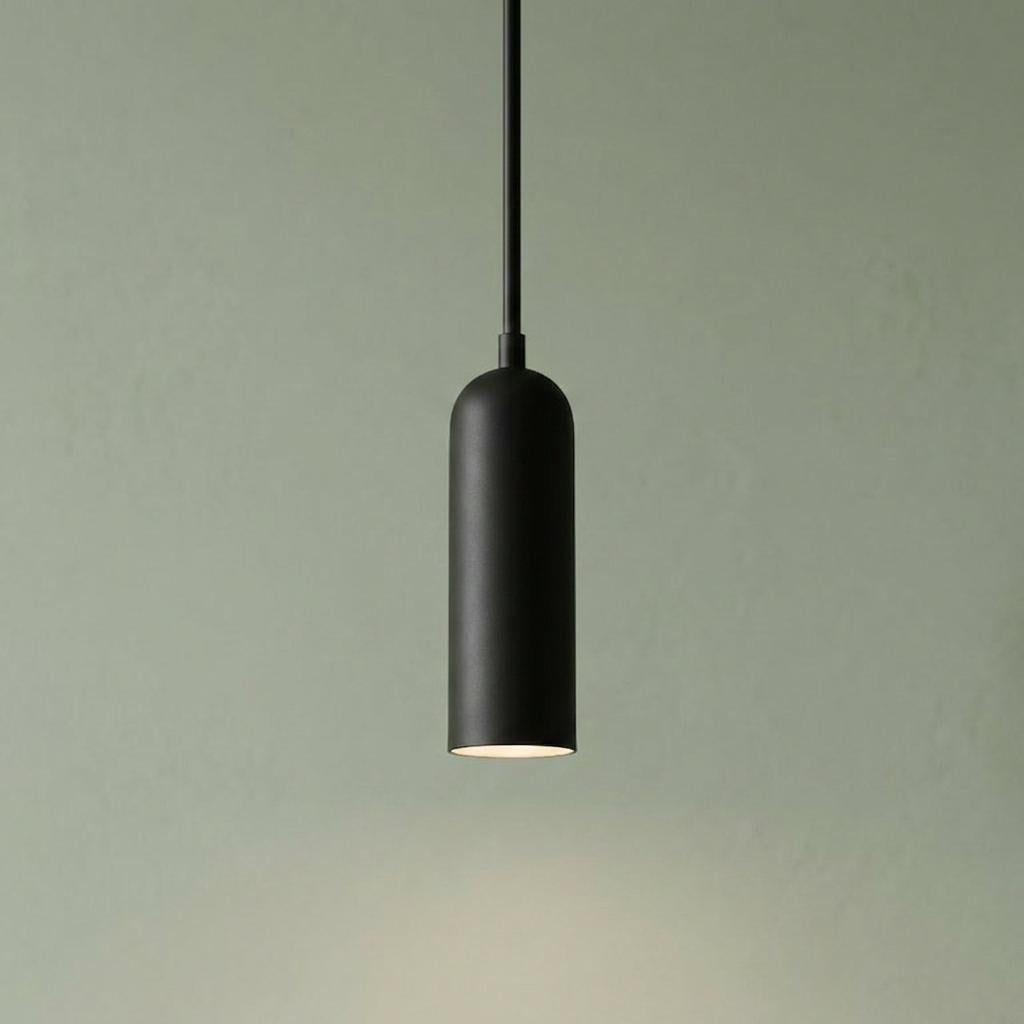 Matte black cylindrical LED mini pendant with a rounded top, emitting warm light against a sage green wall.