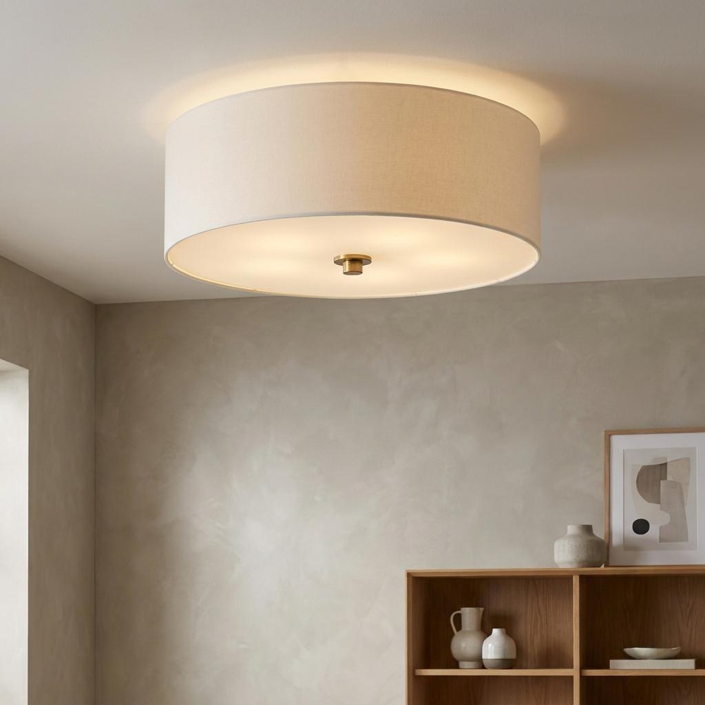 A large drum-shaped linen flush mount ceiling light with a brass finial, glowing in a modern neutral room.
