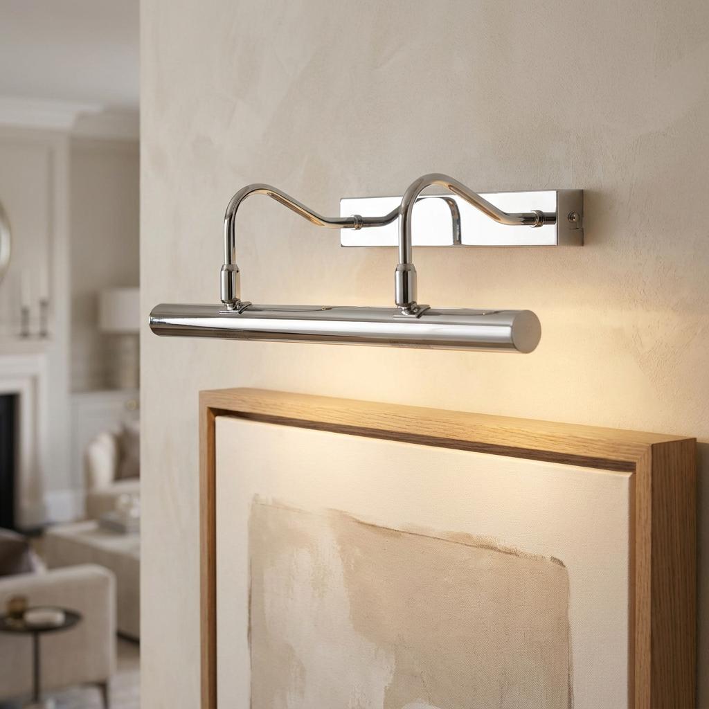 A double arm chrome picture light mounted on a wall, illuminating a framed canvas with a warm glow.