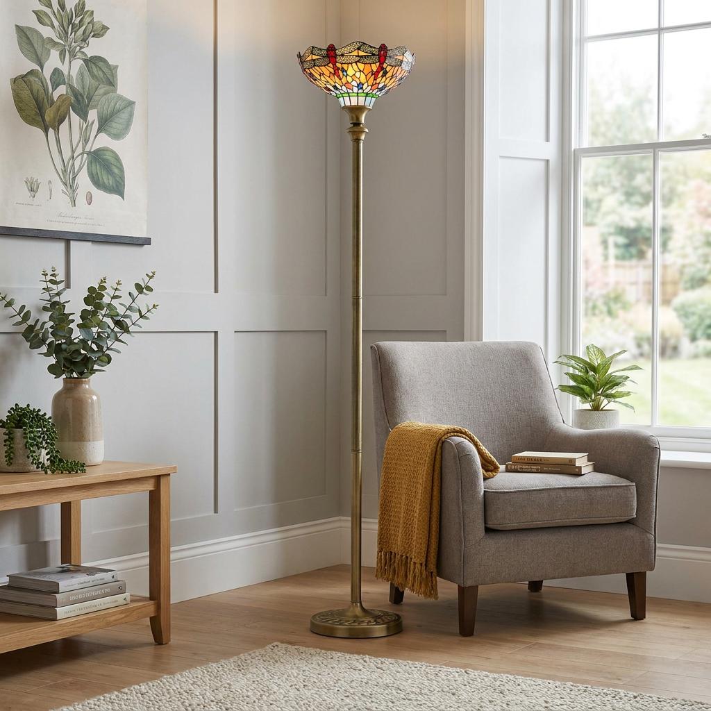 Antique brass floor lamp with a dragonfly stained glass shade standing beside a grey armchair in a living room.