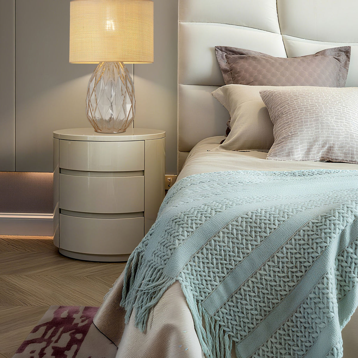 Diane rattan shaded lamp lit on a round bedside chest beside a cream bed with knitted throw