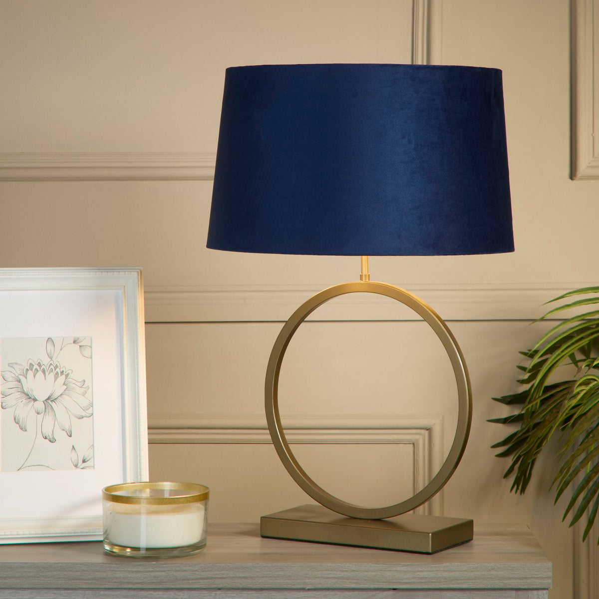 Elegant bronze lamp with navy velvet shade lit on a grey sideboard beside framed art and a candle
