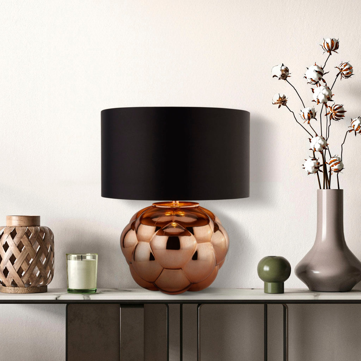 Fizz copper lamp lit on a grey sideboard beside cotton stems in a vase and candle